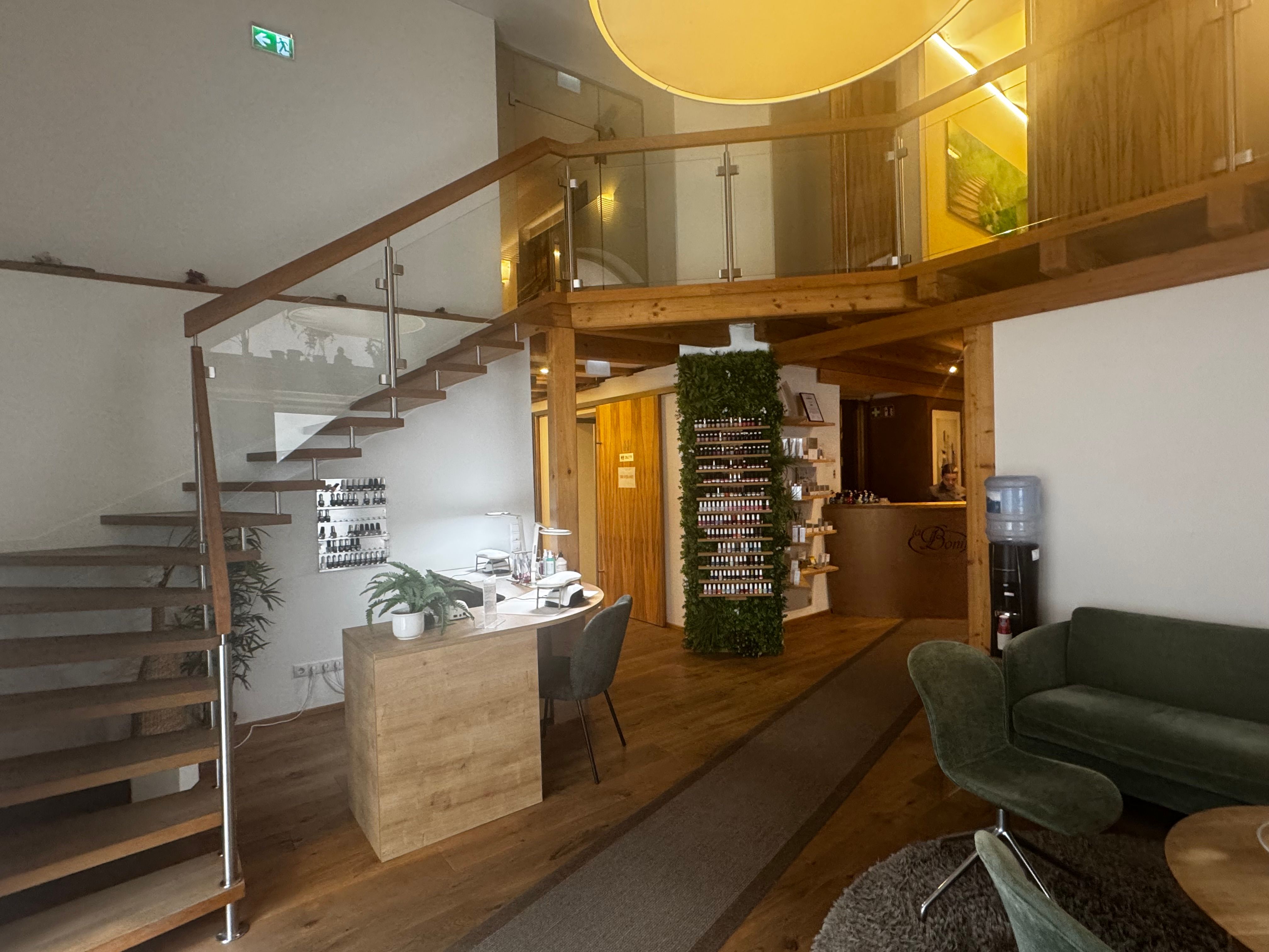 Foto: Innenraum mit offenem Empfangs- und Wartebereich in Holzoptik. Links führt eine Treppe mit Glasgeländer zu einer oberen Galerie. Im Vordergrund steht ein Arbeitsplatz mit Tisch, Stuhl und Pflanzen. In der Mitte befindet sich ein Regal mit kleinen Fläschchen vor einer begrünten Wand. Rechts sind grüne Sitzmöbel und ein Wasserspender 