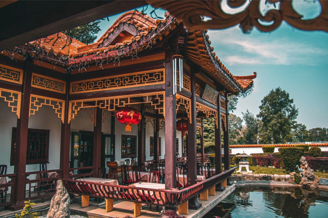 Foto: Ein überdachter Außenbereich von einem Restaurant, der einem chinesischen Tempel ähnelt. Unter dem Dach gibt es Tische und Sessel, davor einen Teich und einen Garten im Hintergrund.