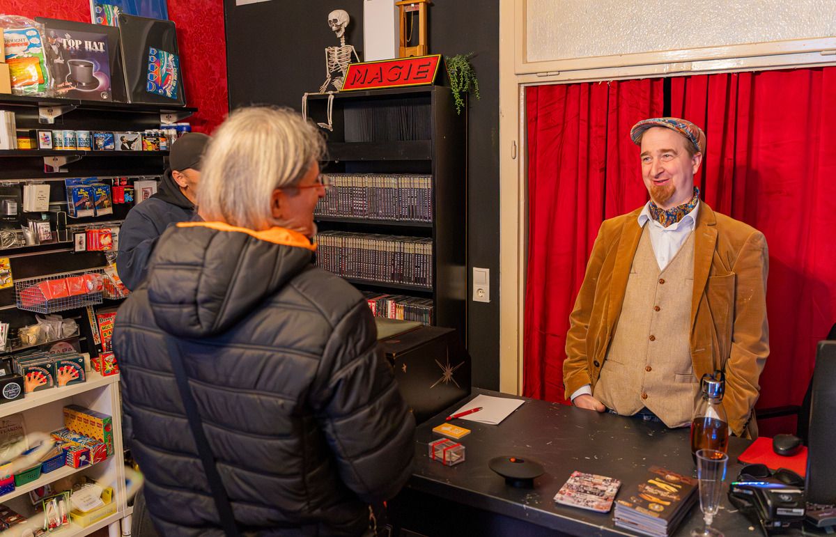 Foto: Ansicht im Geschäft Verkaufspult mit Besitzer und Kunden und vielen Spielen, Tricks und DVDs im Hintergrund. Alle stehen vor einem roten Vorhang