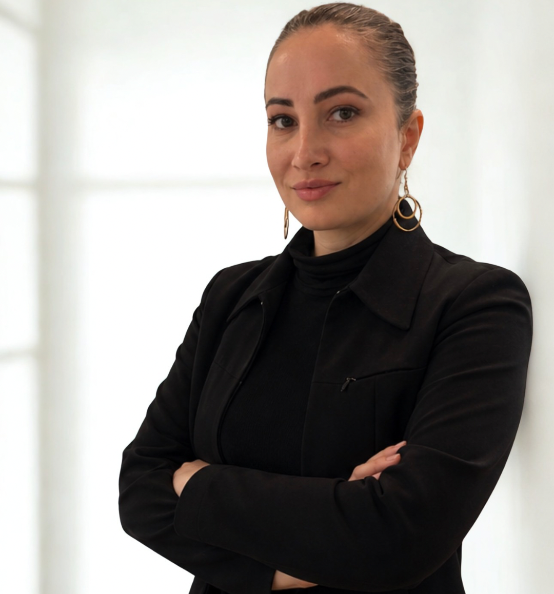 Foto: Porträt einer Person mit zurückgebundenen Haaren und schwarzen Kleidungsstücken. Die Person trägt einen schwarzen Blazer, ein schwarzes Oberteil und große runde Ohrringe. Die Arme sind verschränkt. Der Hintergrund ist hell