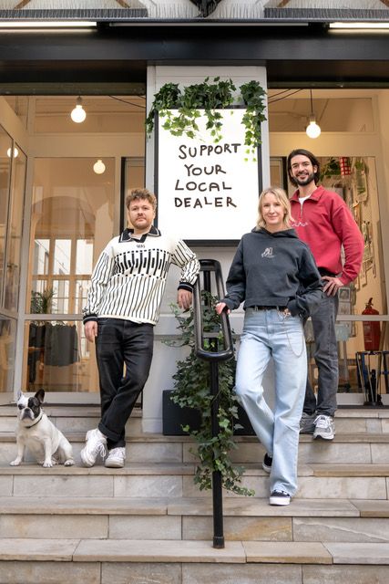 Foto: Porträt von drei Personen und einem Hund, die auf Treppen stehen. Hinter ihnen hängt ein Schild, auf dem unterstütze deinen lokalen Händler steht. Dahinter sind Fensterfronten