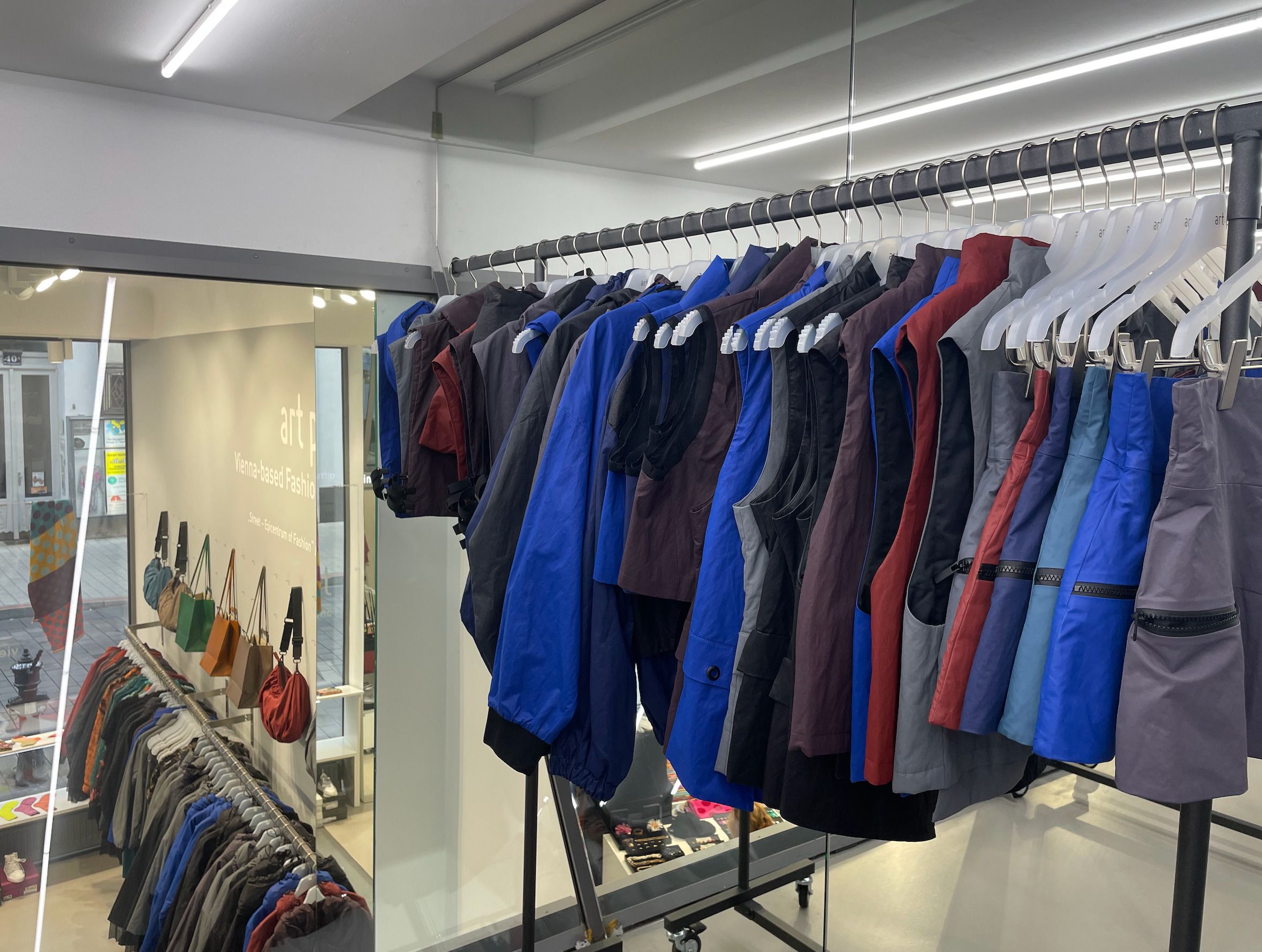 Foto: Rechts ist eine Kleiderstange, auf der verschiedene Oberteile und Hosen hängen. Im Hintergrund ist ein Raum, in dem weitere Kleiderstangen mit Kleidungsstücken und Taschen sind