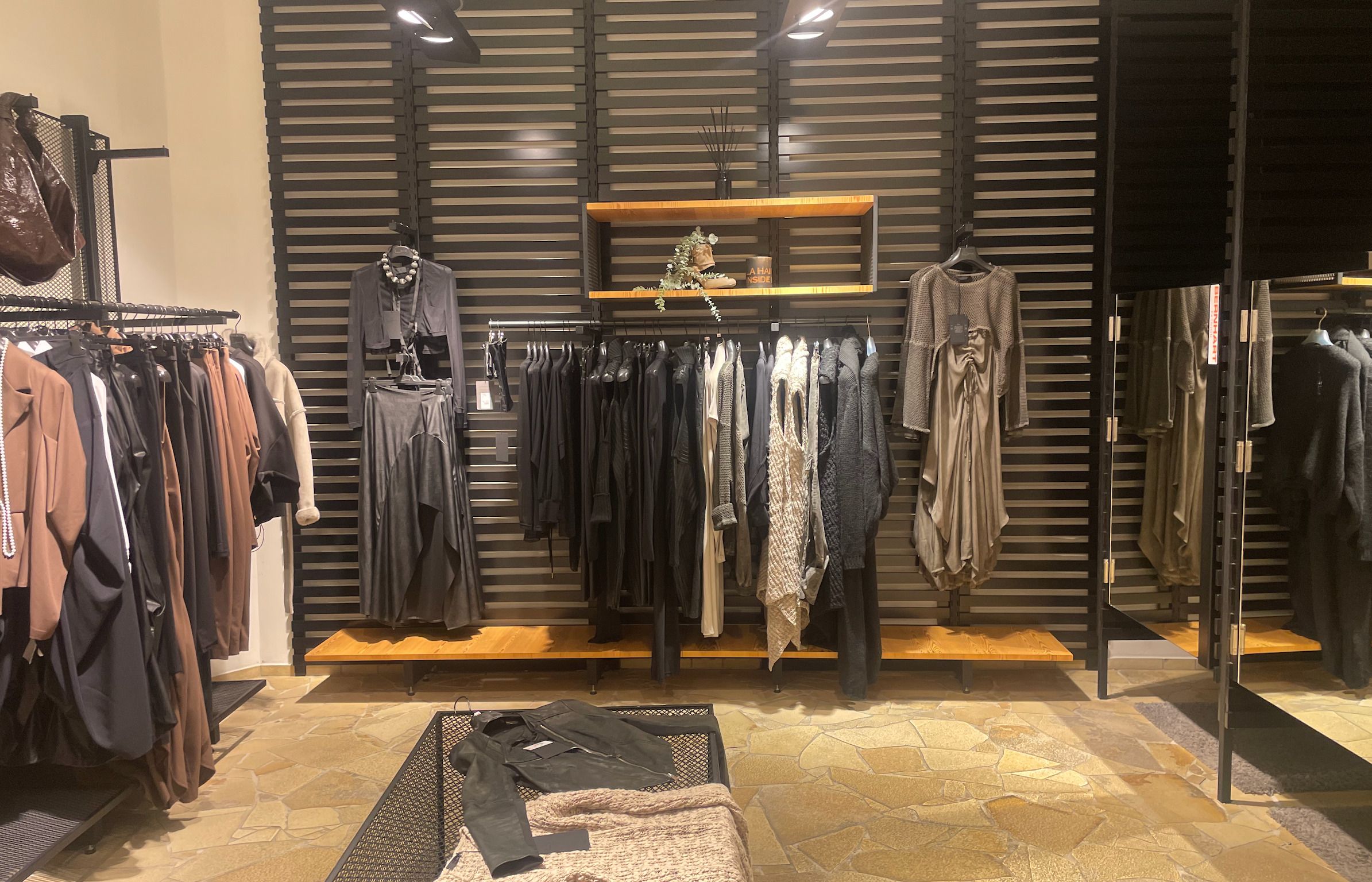 Foto: In einem beleuchteten Raum sind mehrere Kleiderstangen mit verschiedenen Kleidungsstücken. Rechts sind zwei Umkleidekabinen