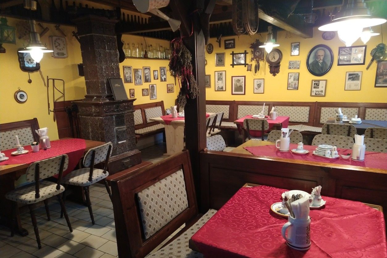 Foto: Gaststube mit Tischen und Stühlen, ein Kamin und eine Wand mit Bildern.