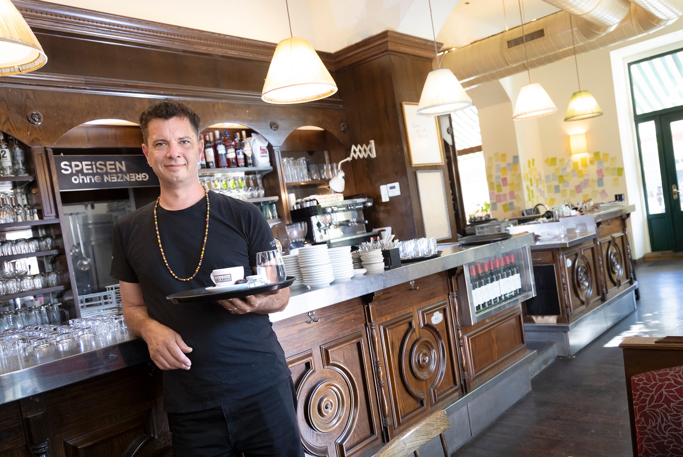 Foto: Innenansicht des Lokals Speisen ohne Grenzen. An der langen Bar steht der Besitzer der freundlich lächelt und einen sehr guten Cafe serviert