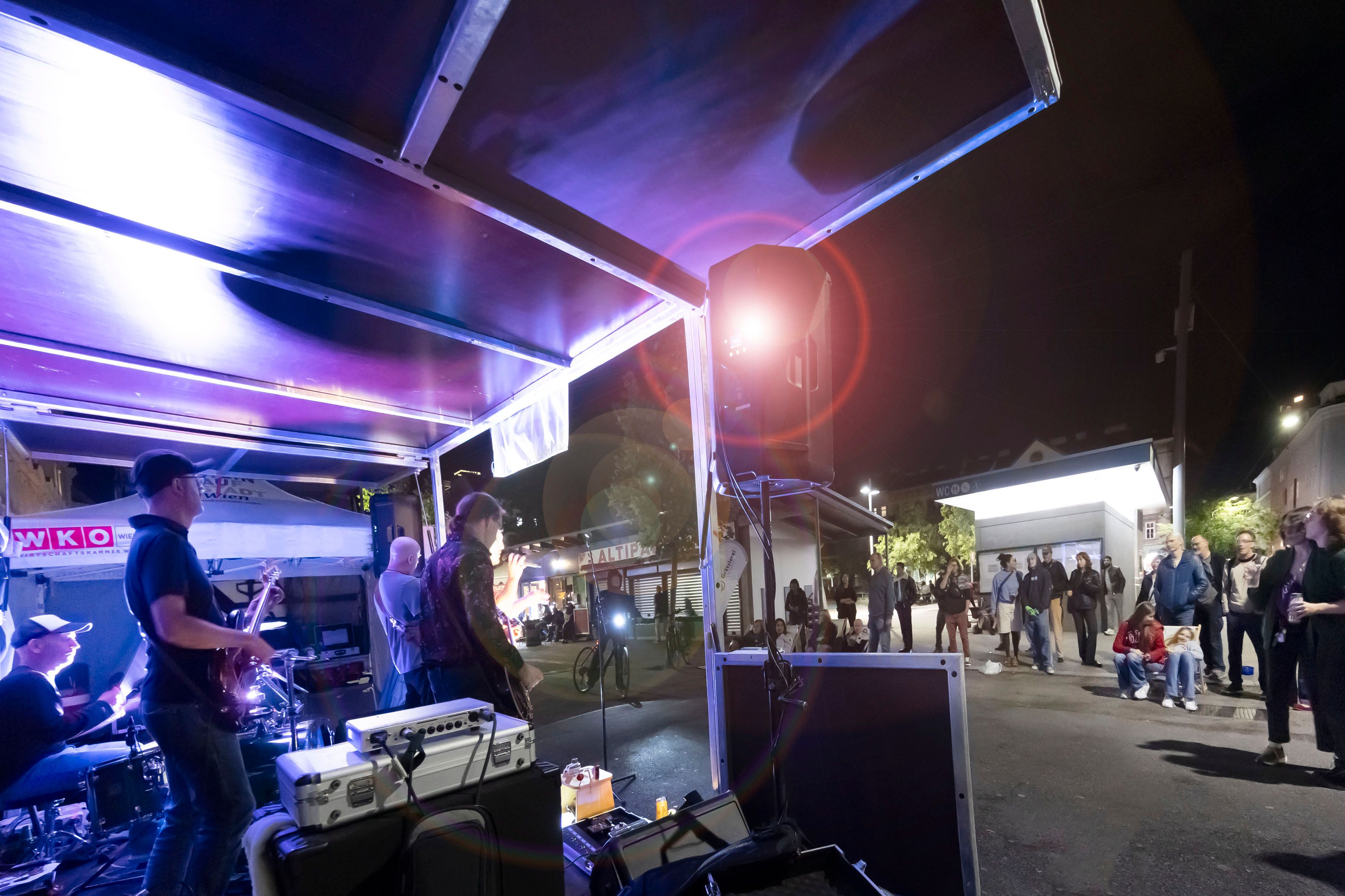 Foto: Bühne mit Musikern aus Seitenansicht am Abend mit blauem Licht und einigen Leuten vor der Bühne.