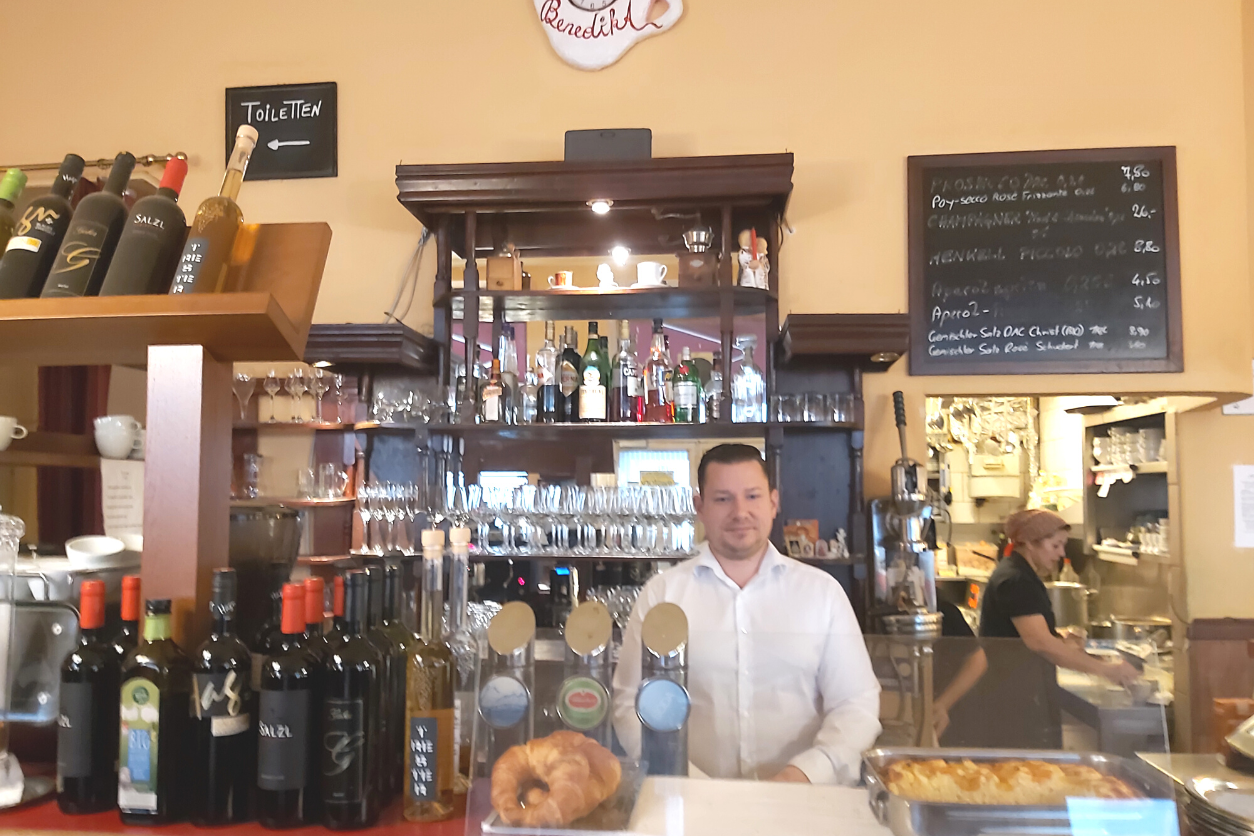 Foto: Zeigt einen Mann hinter einer Bar mit Weinflaschen und Croissants