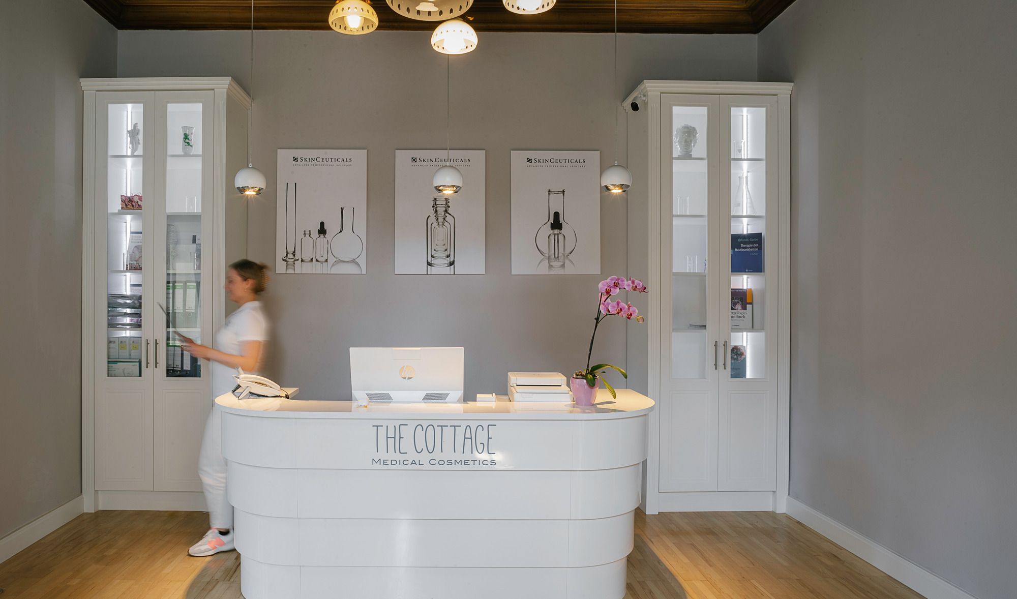 Foto: Ein weißer Empfangstresen, auf dem steht „The Cottage Medical Cosmetics“. Hinter dem Empfang hängen drei gerahmte Bilder mit Laborgefäßen und dem Schriftzug „SkinCeuticals“. Links und rechts befinden sich hohe weiße Vitrinenschränke. Eine Person in weißer Kleidung ist hinter dem Tresen