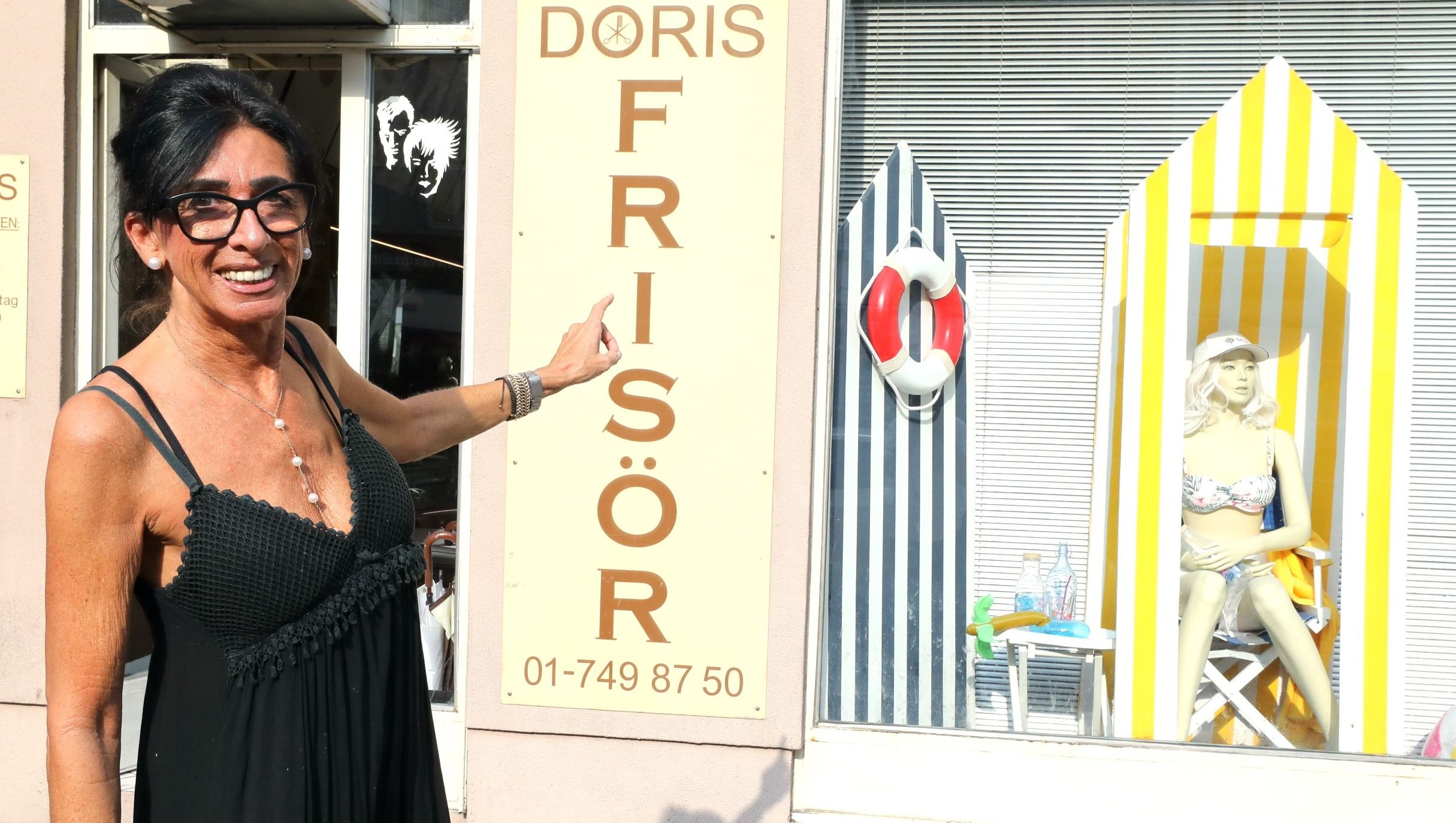 Foto: Eingangsbereich des Frisörsalons, Dame zeigt auf die sommerlich beschmückte Auslage.