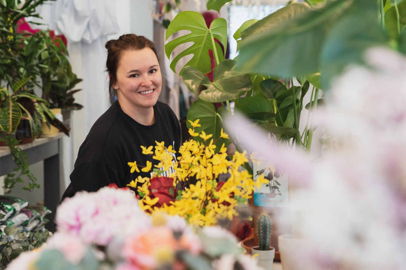 Foto: Blumenverena sitzt mit dunklem T-Shirt hinter bunten Blumen und lächelt freundlich