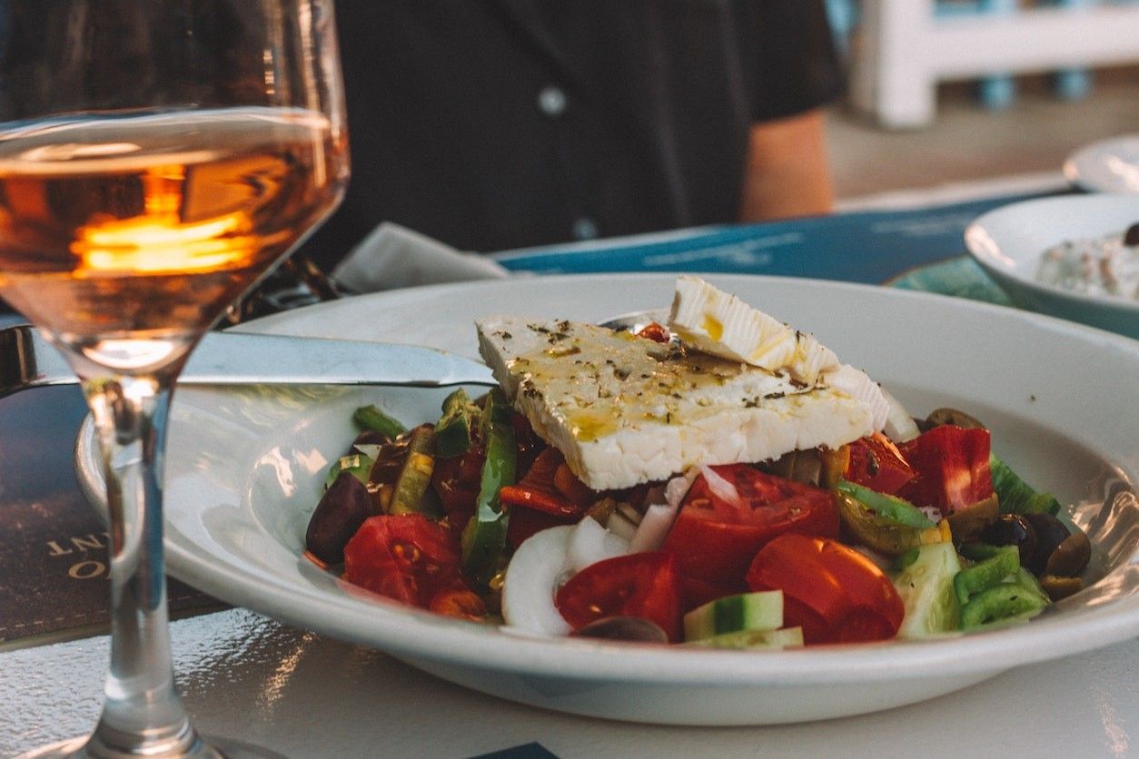 Foto: Ein griechischer Salat mit Feta auf einem Teller. Daneben steht ein Glas Wein.