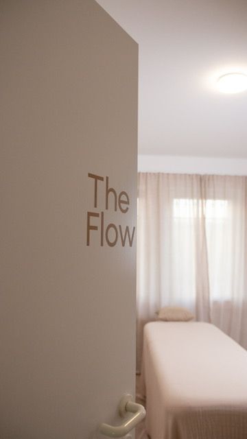 Foto:  Geöffnete Tür mit dem Schriftzug „The Flow“ in brauner Schrift auf heller Oberfläche. Im Hintergrund befindet sich ein Raum mit weiß bezogener Liege